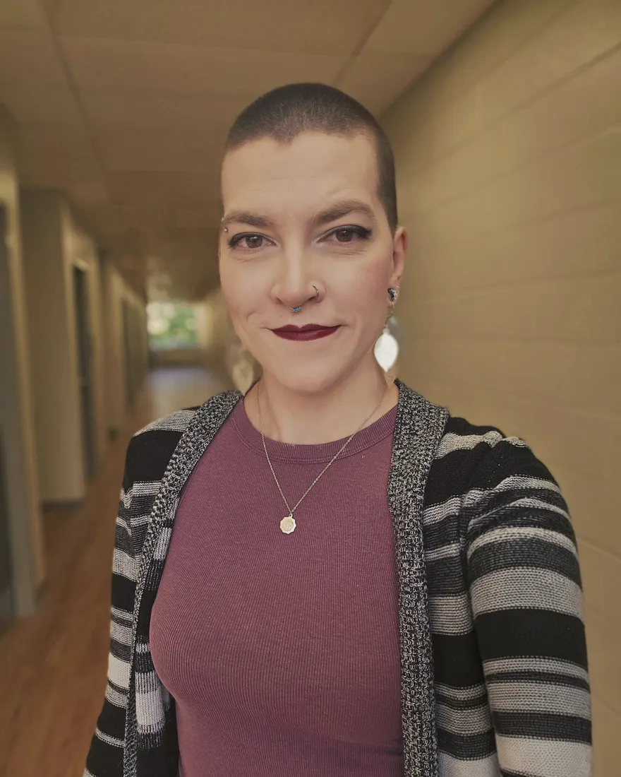 Young woman with short dark hair and dark red lipstick, smiling, while standing in an office hallway.