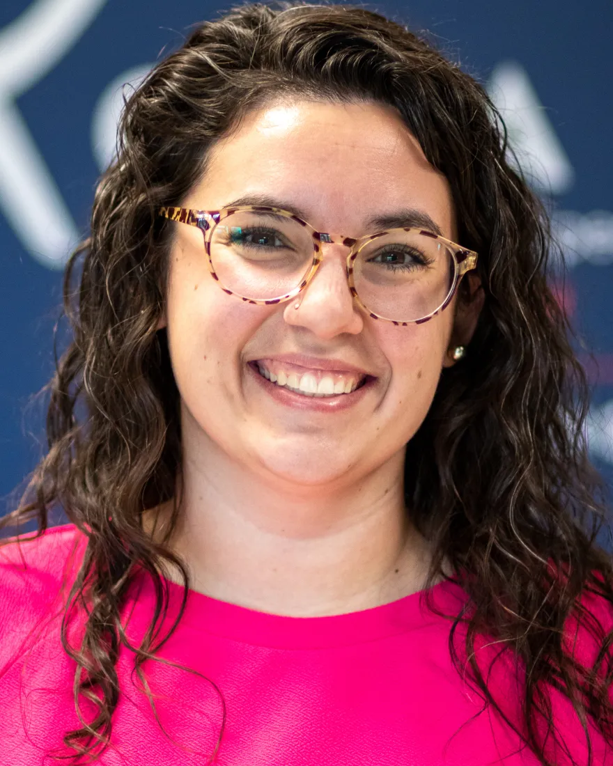 Headshot of Natalie Simpson, with shoulder length brown curly hair, glasses, and a pink shirt against a navy backdrop.