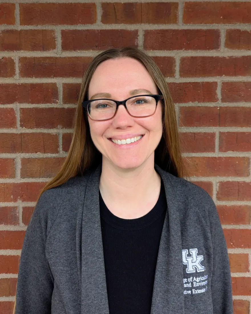 A woman with long blonde hair and glasses stands smiling in front of a brick wall, wearing a dark top with a University of Kentucky logo.