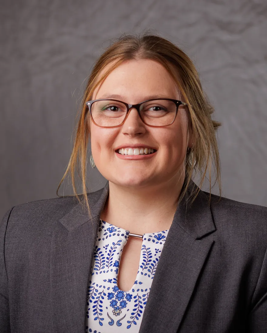 This is a professional headshot of a woman smiling at the camera. She is dressed in a gray blazer over a white blouse with a blue floral pattern. The background is a neutral gray backdrop.